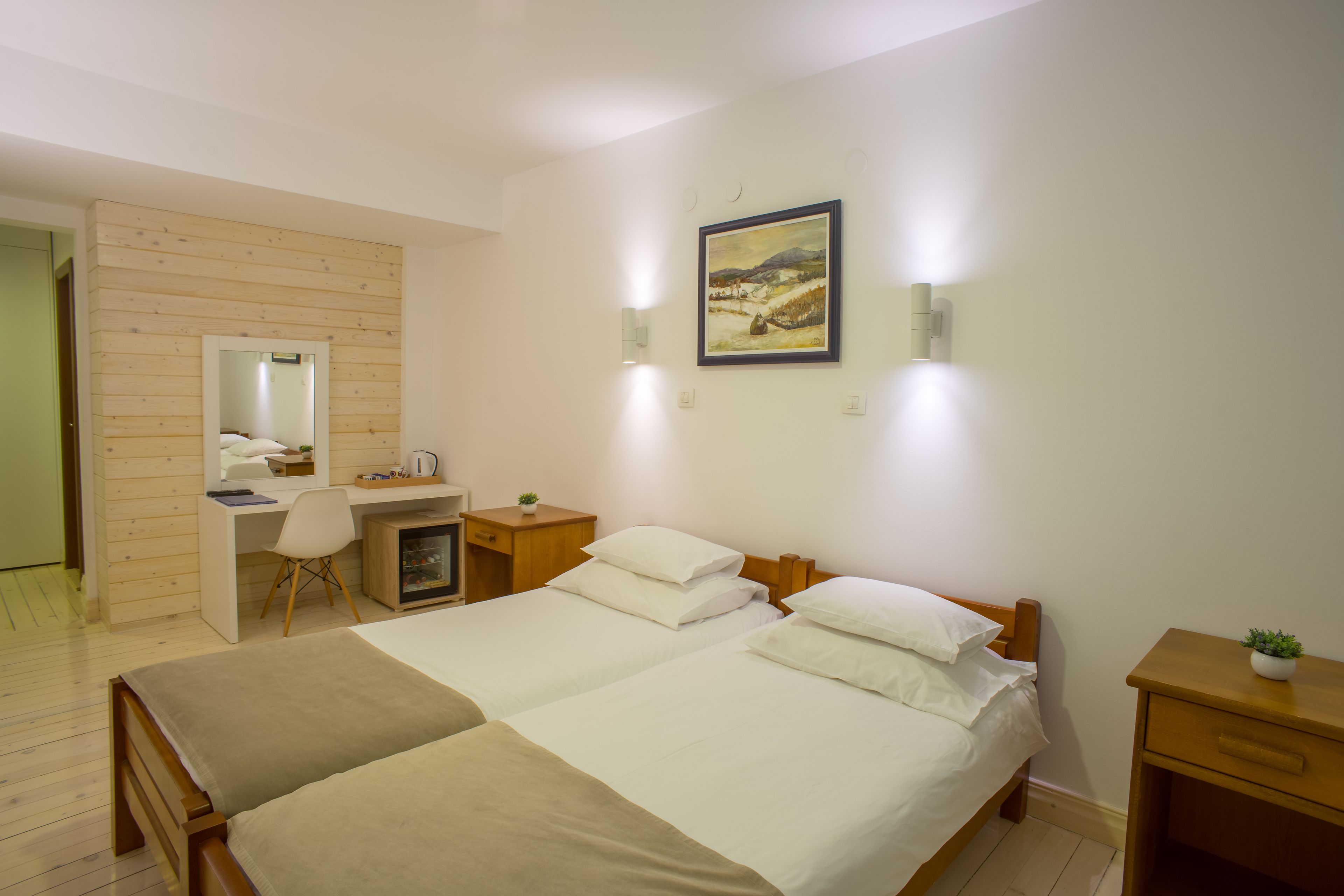 A bedroom with a wooden desk, two beds, a mirror, and a painting on the wall.