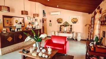 A reception area with a wooden desk, a red chair, and a person at the desk. There are framed pictures on the wall, a large mirror, and a staircase to the right.