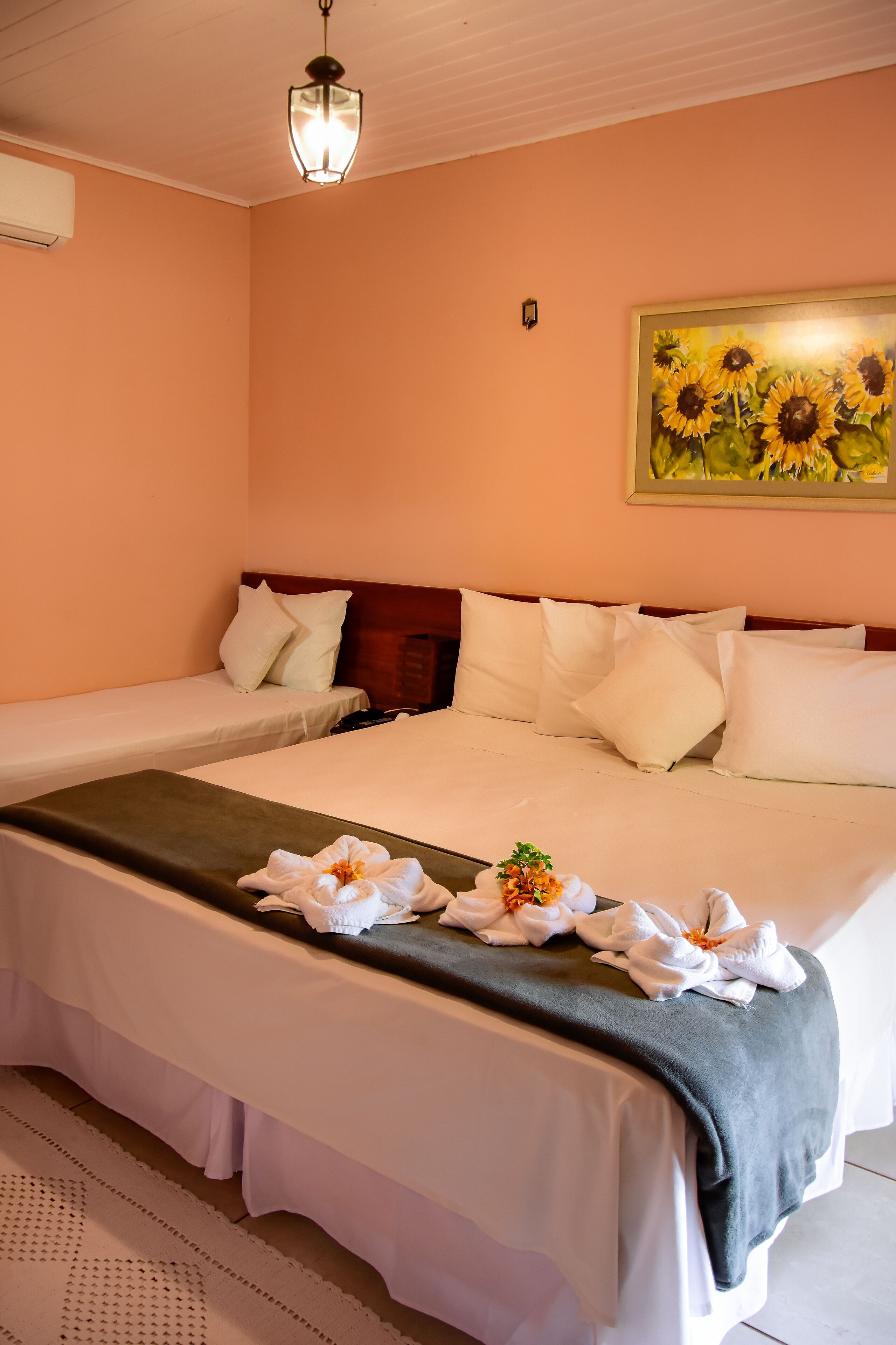 A hotel room with two single beds, a large bed, a painting of sunflowers, a ceiling light, and a wall-mounted air conditioner.