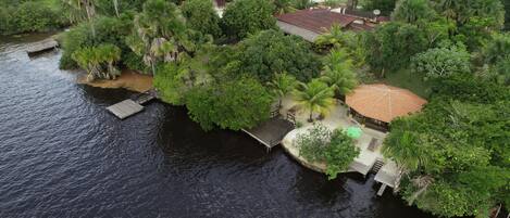 An aerial view of a riverside property with a wooden dock, a small pavilion, and lush greenery.