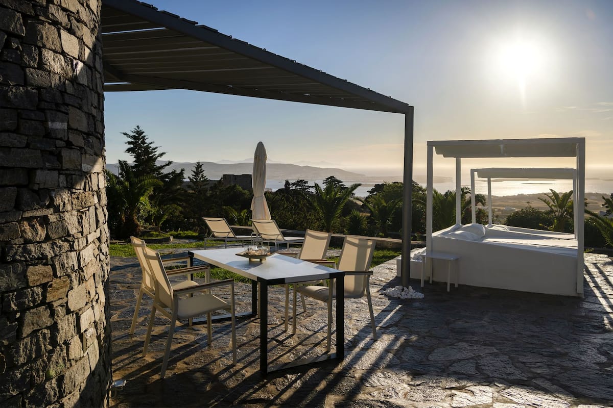 Un área de estar al aire libre con una mesa y sillas, una zona cubierta para sentarse, y vistas al mar y a las montañas.