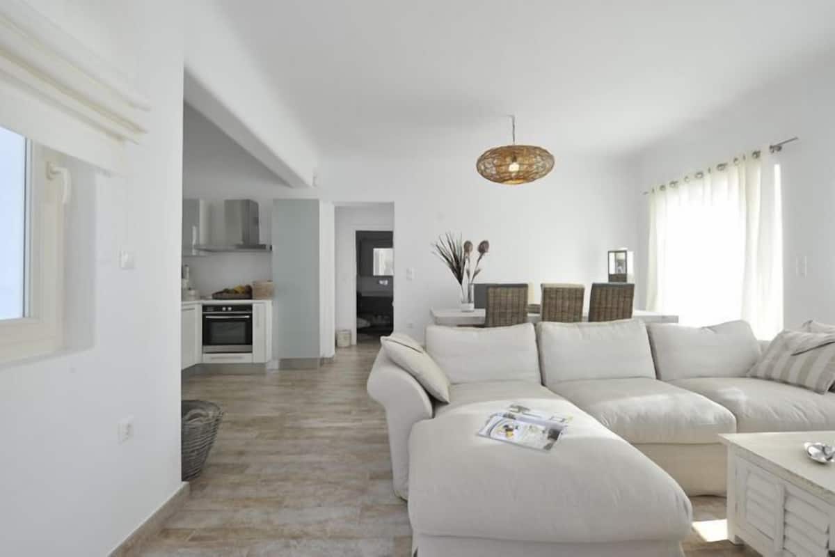 Una sala moderna con un sofá beige, una mesita y una cesta de mimbre. Se observa una cocina al fondo con armarios blancos y una lámpara colgante.
