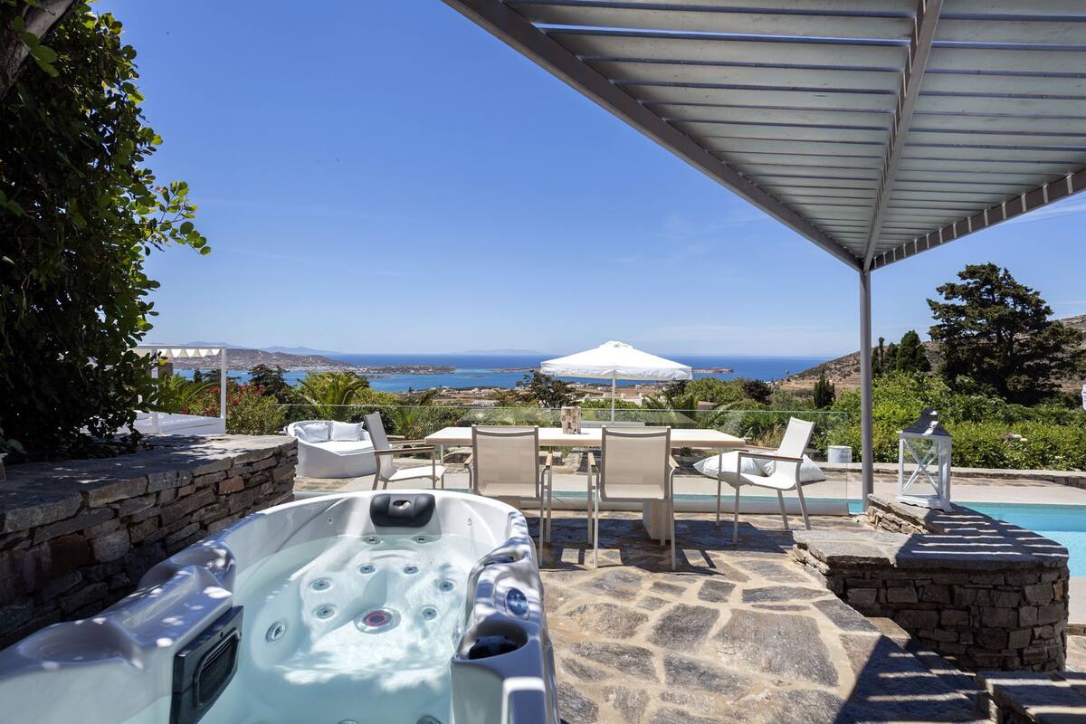 Un jacuzzi con un muro de piedra de fondo, un porche cubierto con mobiliario exterior y vistas al mar y al paisaje distante.
