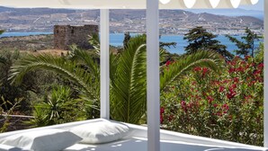 Habitació amb llit, dosser blanc i vista a un paisatge costaner que inclou un castell, vegetació i una massa d'aigua.