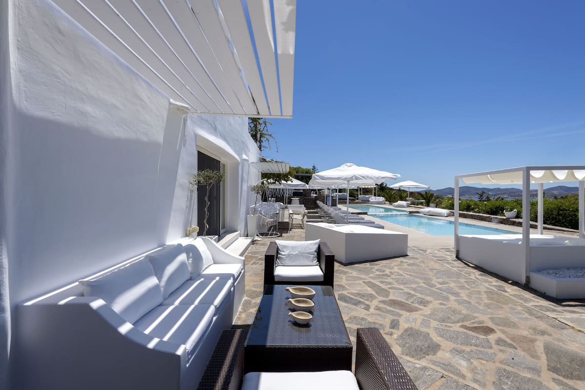Un área junto a la piscina con mobiliario blanco, suelo de piedra y sombrillas.