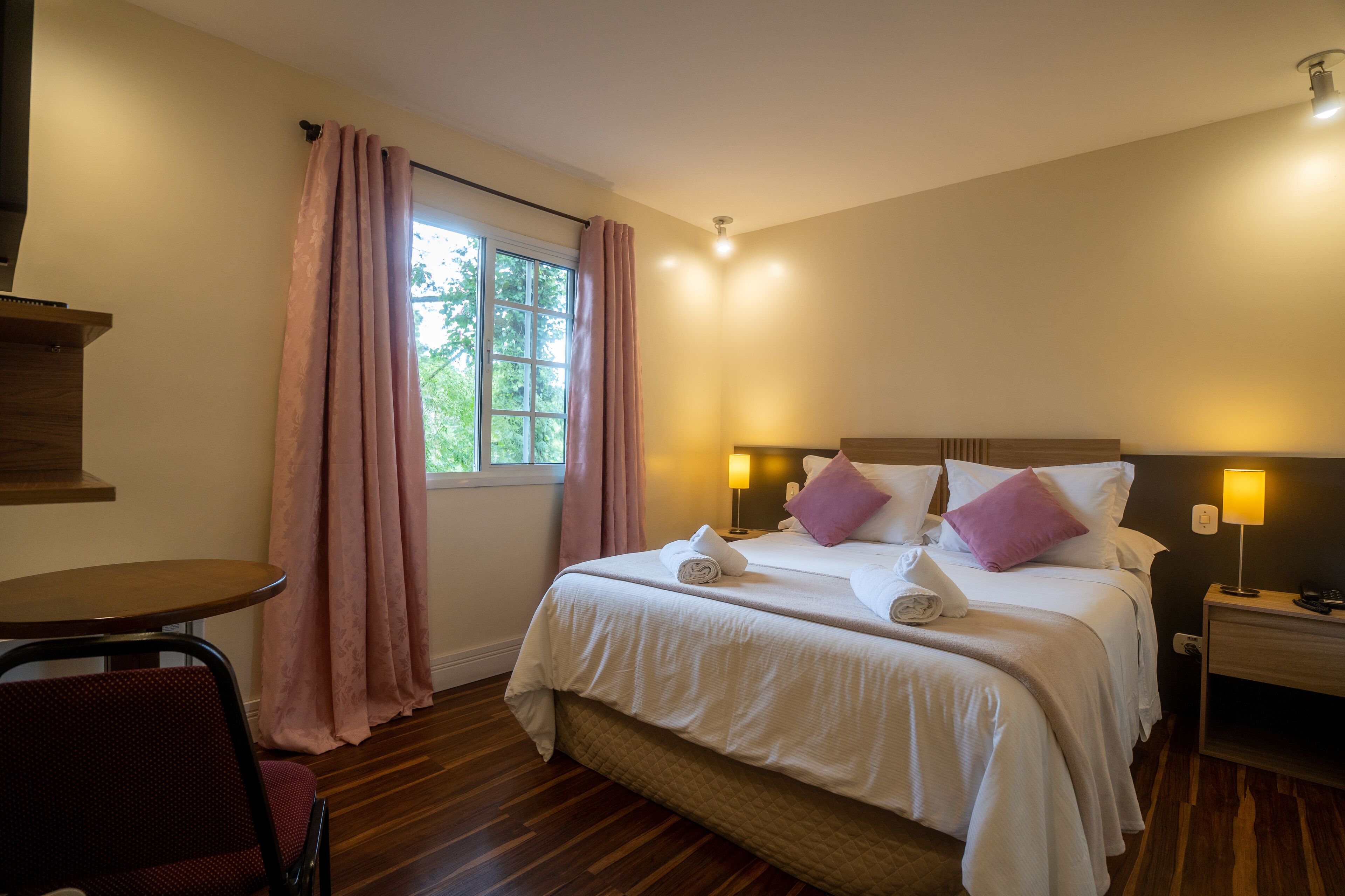 Une chambre d’hôtel avec un lit, des tables de chevet, une chaise, une fenêtre avec des rideaux et un plancher en bois.