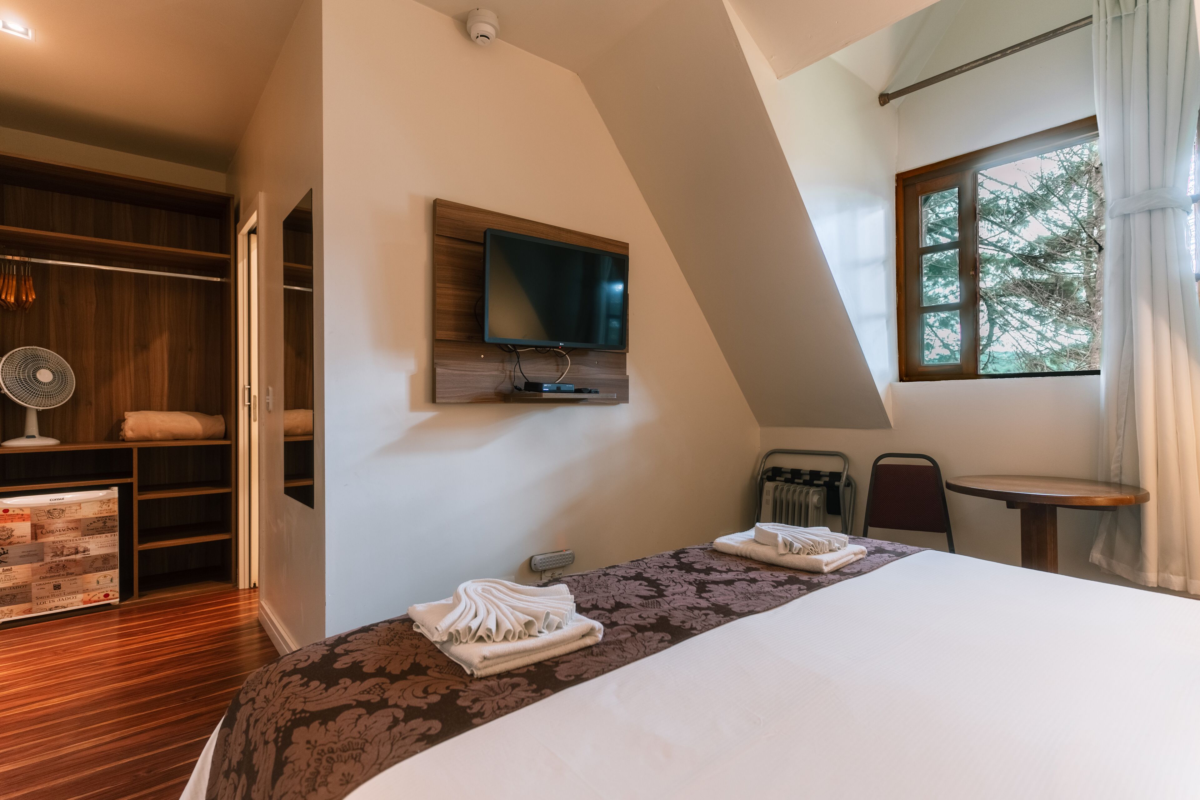 A bedroom with a large bed, a TV mounted on the wall, a wooden wardrobe, and a window with curtains.