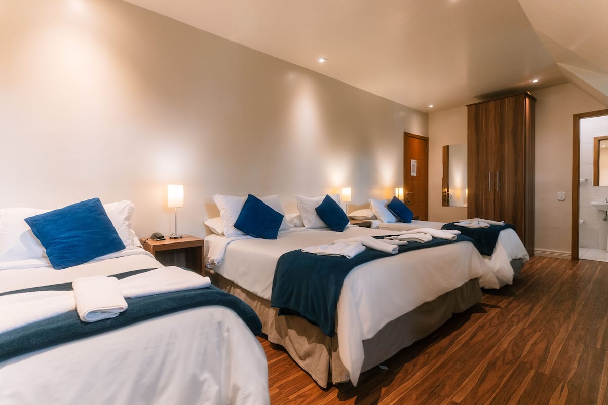 Hotellihuone, jossa on kolme sänkyä, jokaisessa valkoiset lakanat ja siniset tyynyt, puinen vaatekaappi sekä kylpyhuone, jossa on peili ja pesuallas.