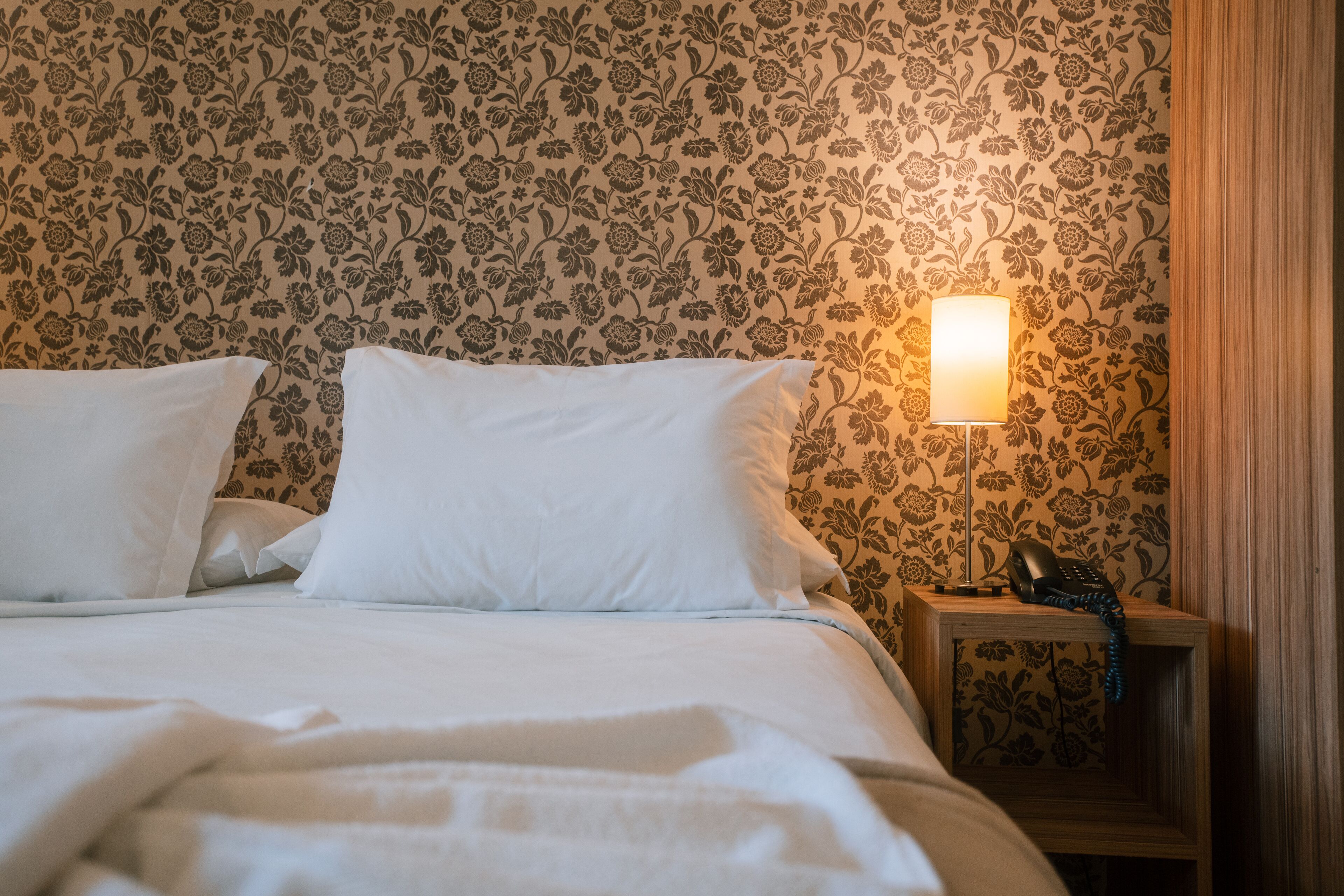 A neatly made bed with white bedding and pillows, a bedside lamp, and a telephone on a wooden nightstand.