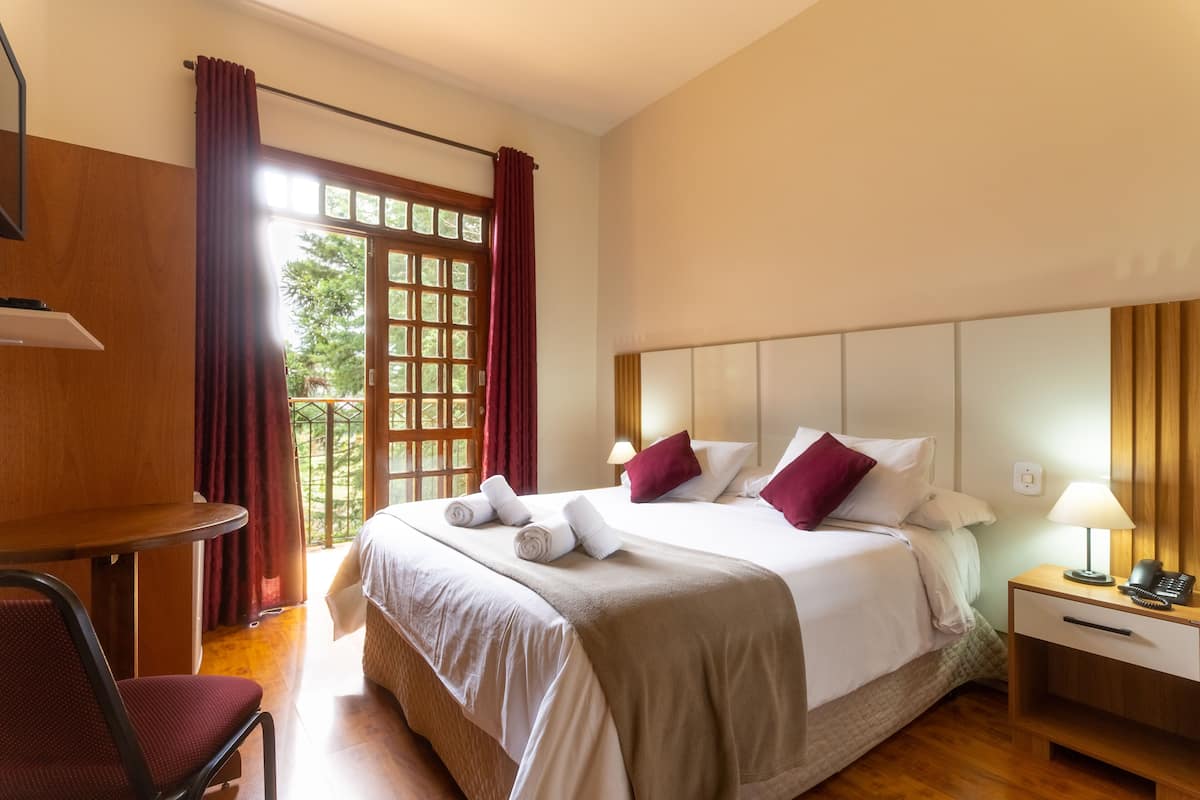 A hotel room with a bed, a desk, a chair, a nightstand with a lamp, and a window with curtains.
