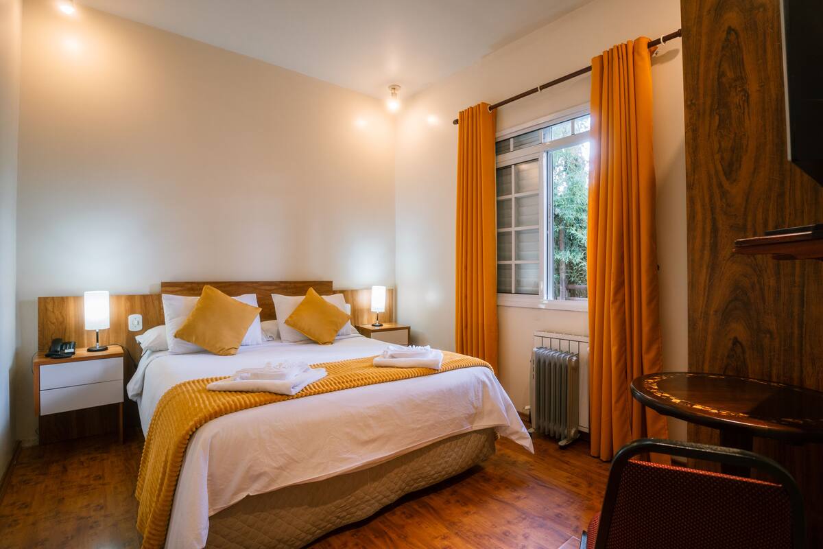 Hotellihuone, jossa on sänky, yöpöydät, tuoli, ikkuna verhoilla ja patteri.