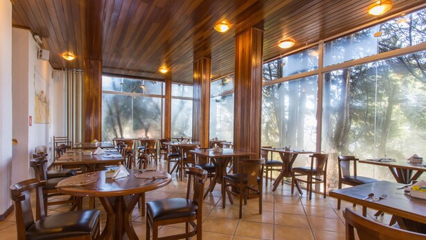 A spacious dining area with wooden tables and chairs, large windows offering a view of trees, and a high ceiling with wooden beams. - Pousada La Toscana (Campos do Jordão)