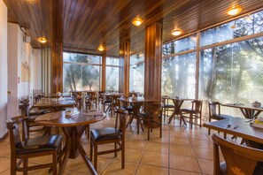 A spacious dining area with wooden tables and chairs, large windows offering a view of trees, and a high ceiling with wooden beams. - Pousada La Toscana (Campos do Jordão)