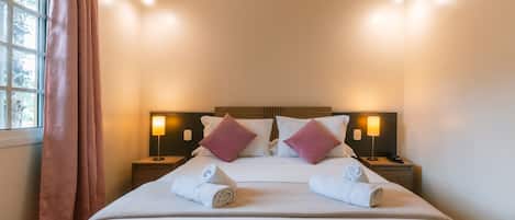 A neatly made bed with white linens and two pink pillows, a window with curtains, and two bedside lamps.