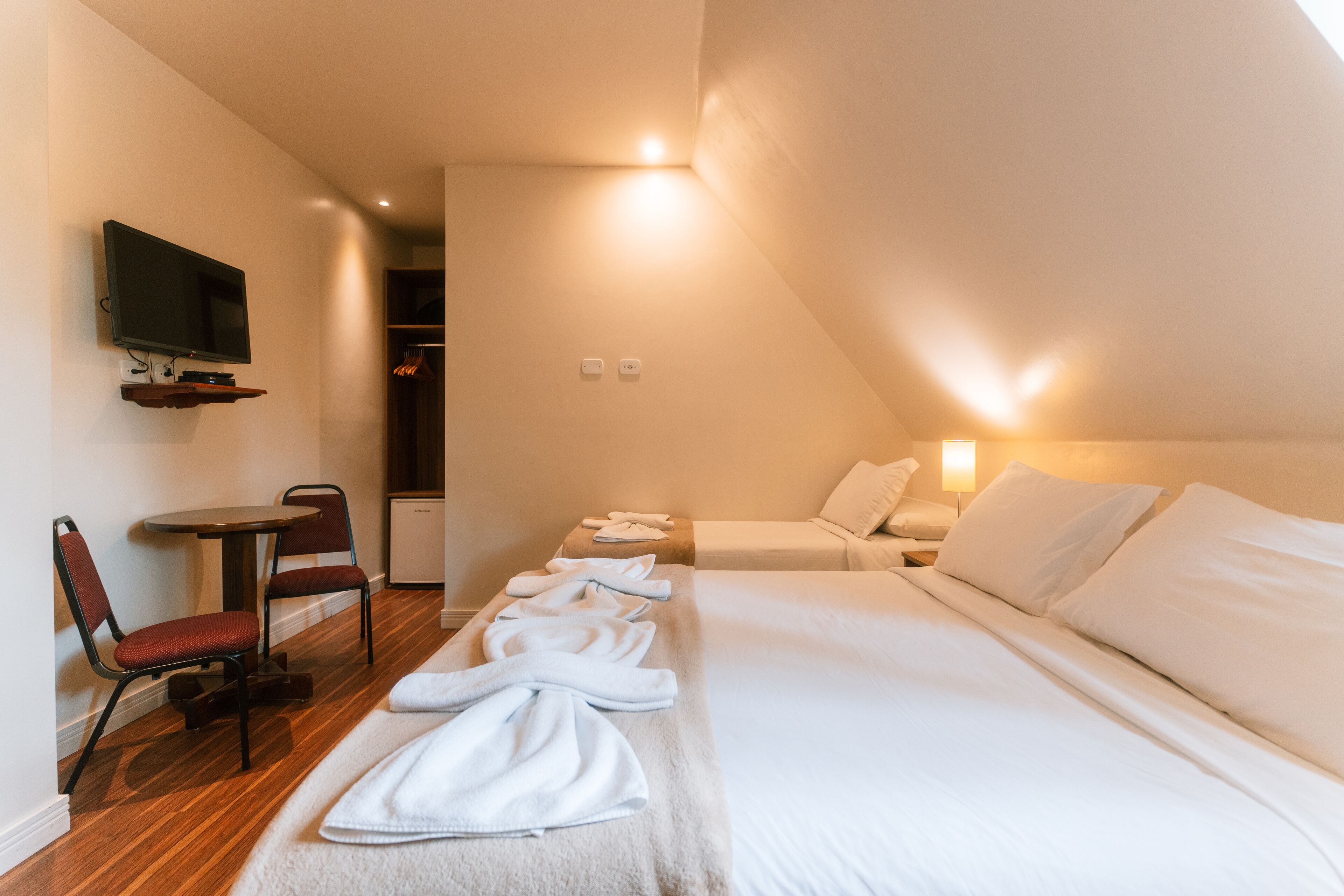 A hotel room with a bed, a TV mounted on the wall, a small table with chairs, and a closet.