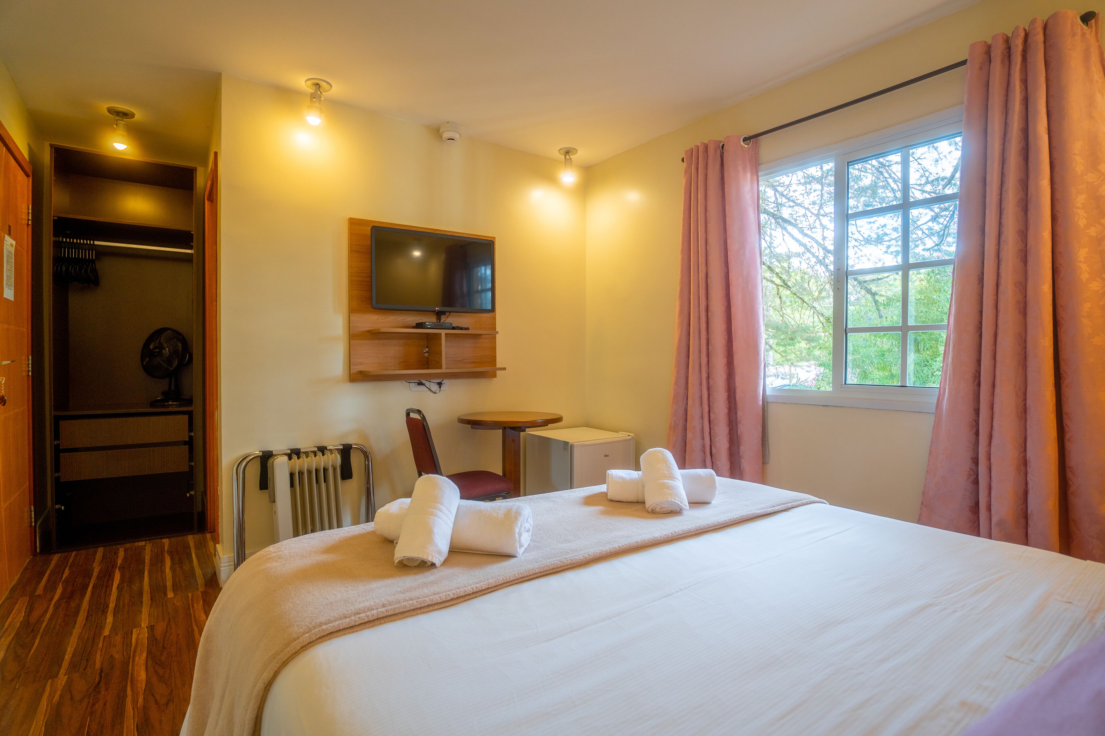 A hotel room with a bed, a TV mounted on the wall, a desk, a chair, and a window with curtains.