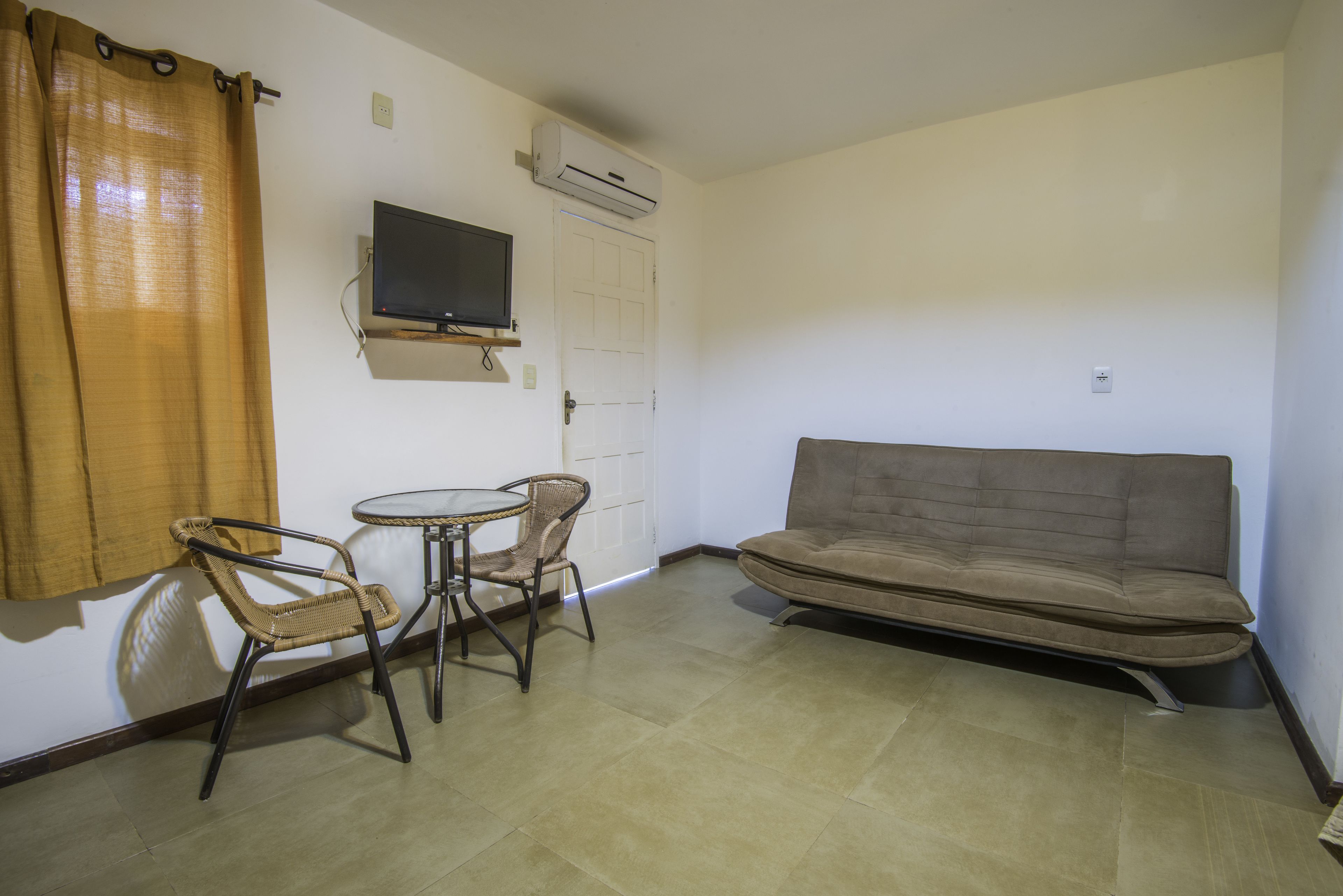 Quarto com sofá, duas cadeiras, uma pequena mesa, televisão fixada na parede e aparelho de ar condicionado.