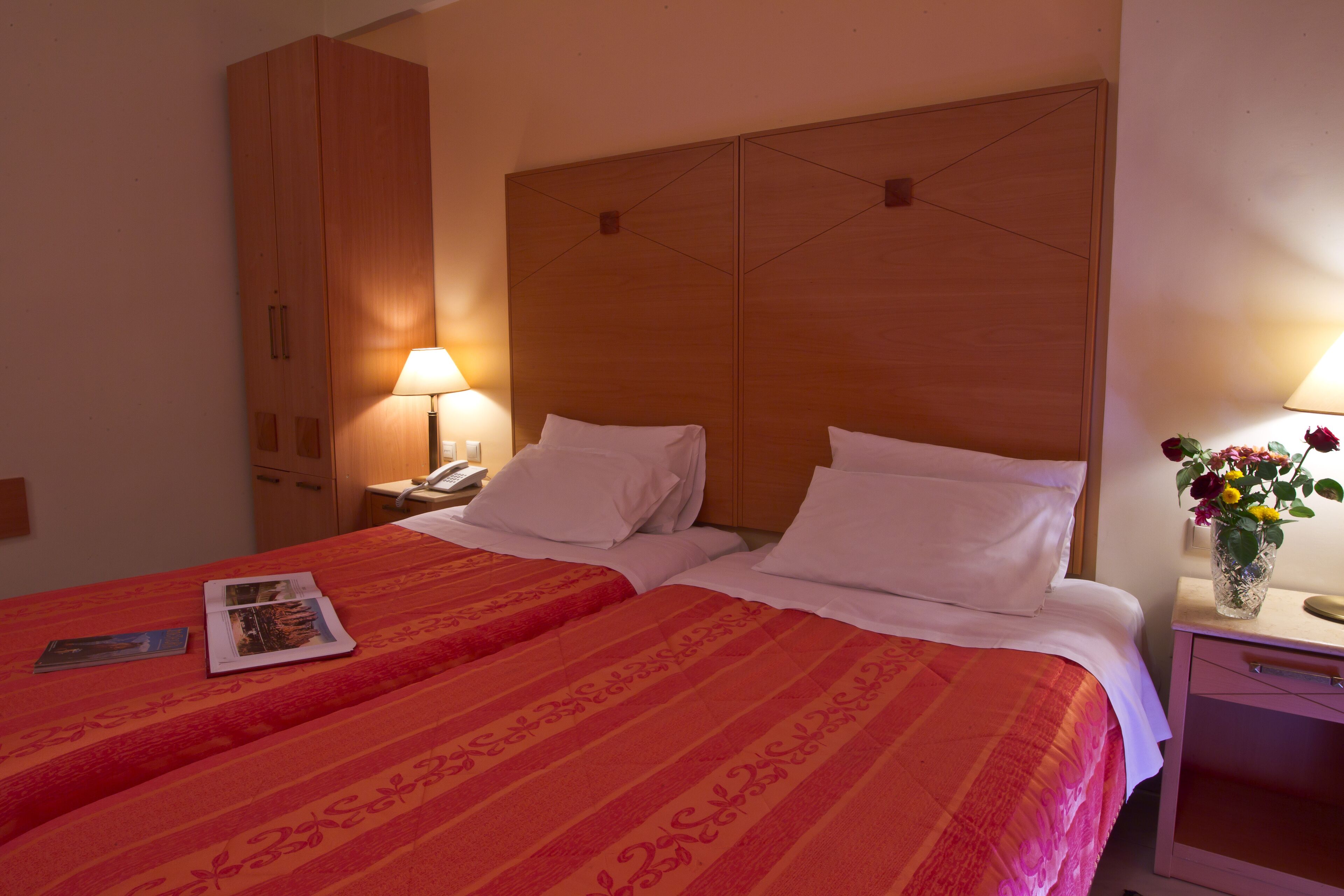 A hotel room with two beds, a wooden wardrobe, a bedside table with a lamp, and a vase with flowers.