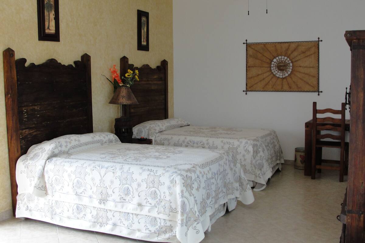 Un dormitorio con dos camas, un ventilador de techo, una silla de madera y un adorno en la pared.