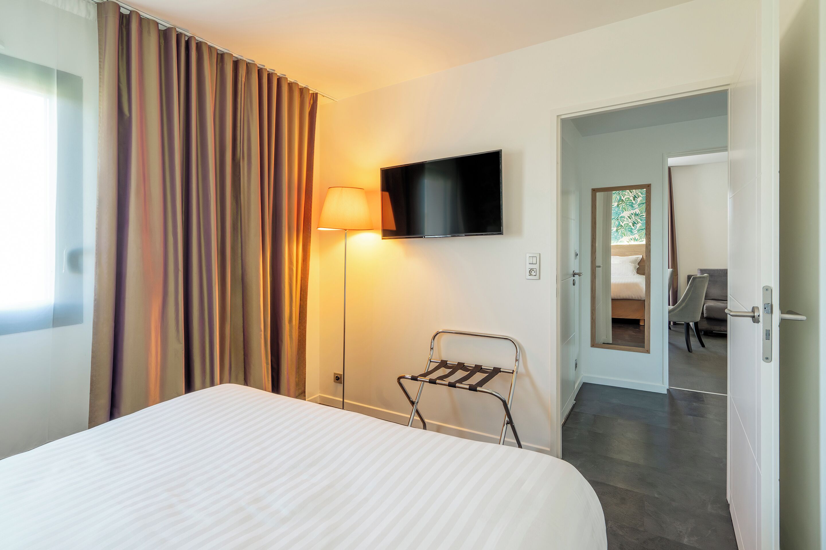 Ein Hotelzimmer mit Bett, Wandfernseher, Lampe und Blick auf ein weiteres Zimmer durch eine offene Tür.