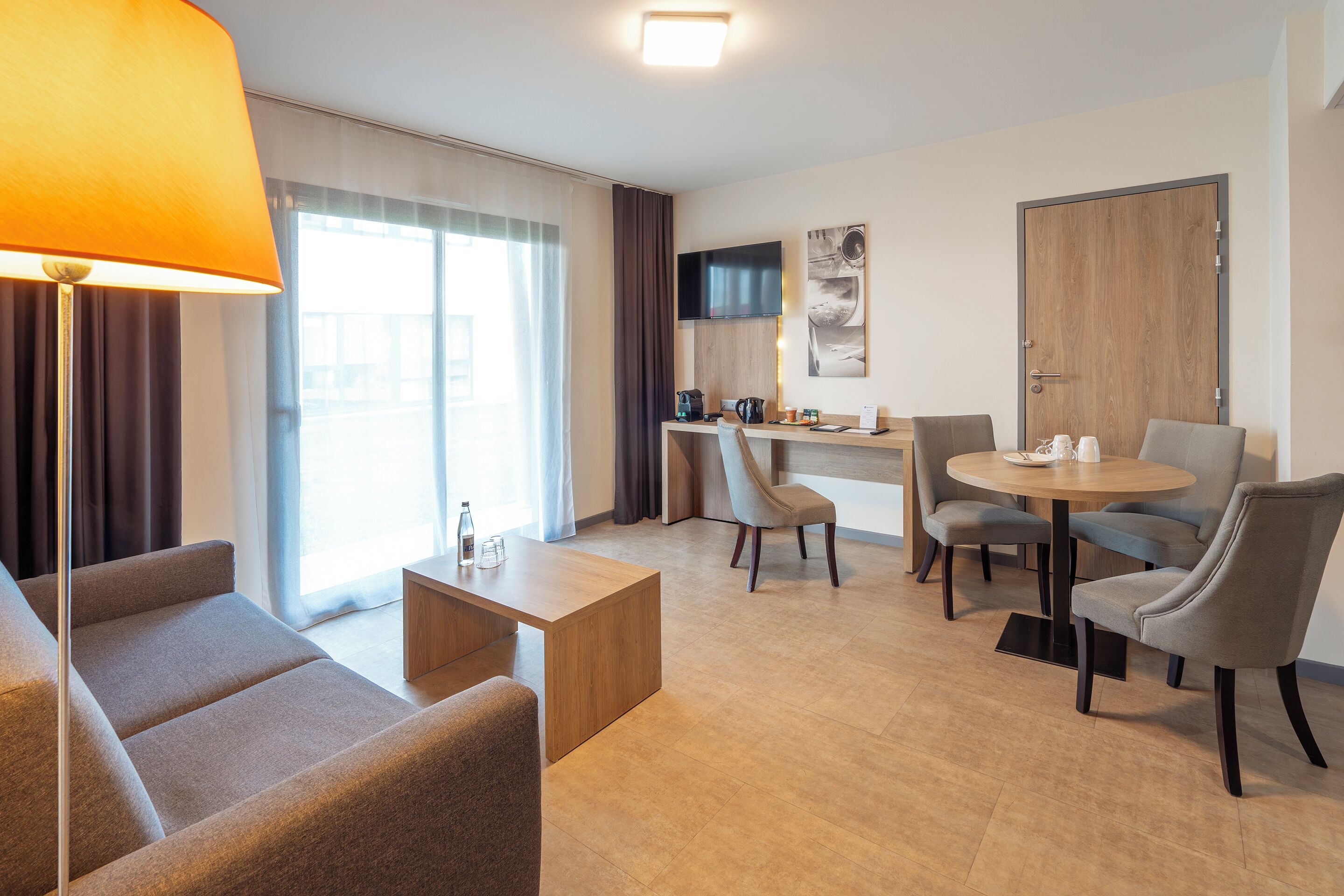 Une chambre d’hôtel moderne équipée d’un canapé, d’une petite table, d’une table de salle à manger ronde avec des chaises, d’un coin cuisine et d’une grande fenêtre avec des rideaux.