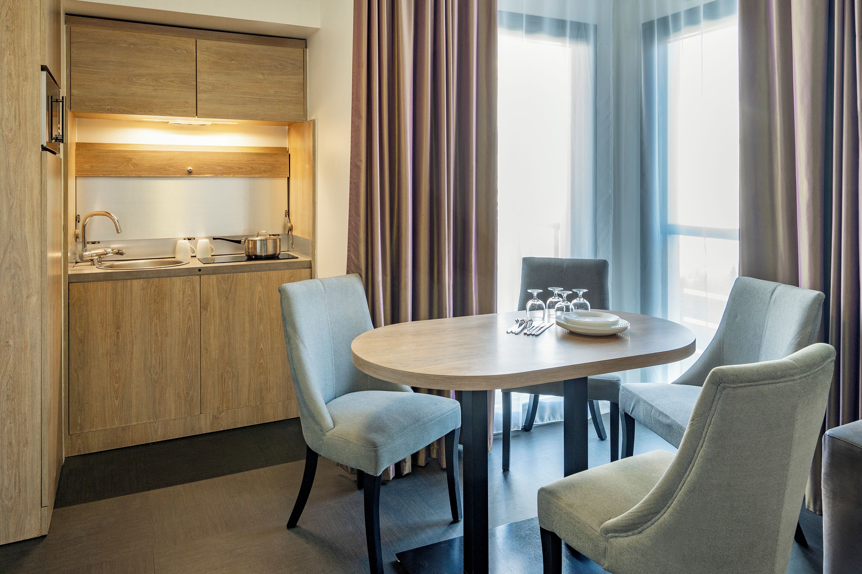 A modern hotel room with a wooden kitchenette, a round dining table with four chairs, and large windows with curtains.