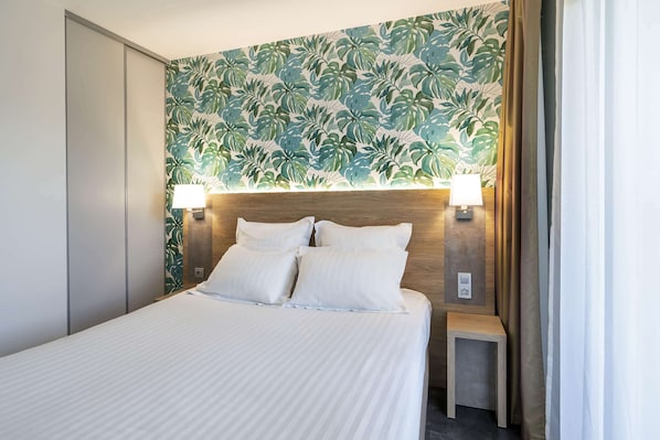 A hotel room with a bed, two bedside lamps, a nightstand, and a wall with a tropical leaf pattern.