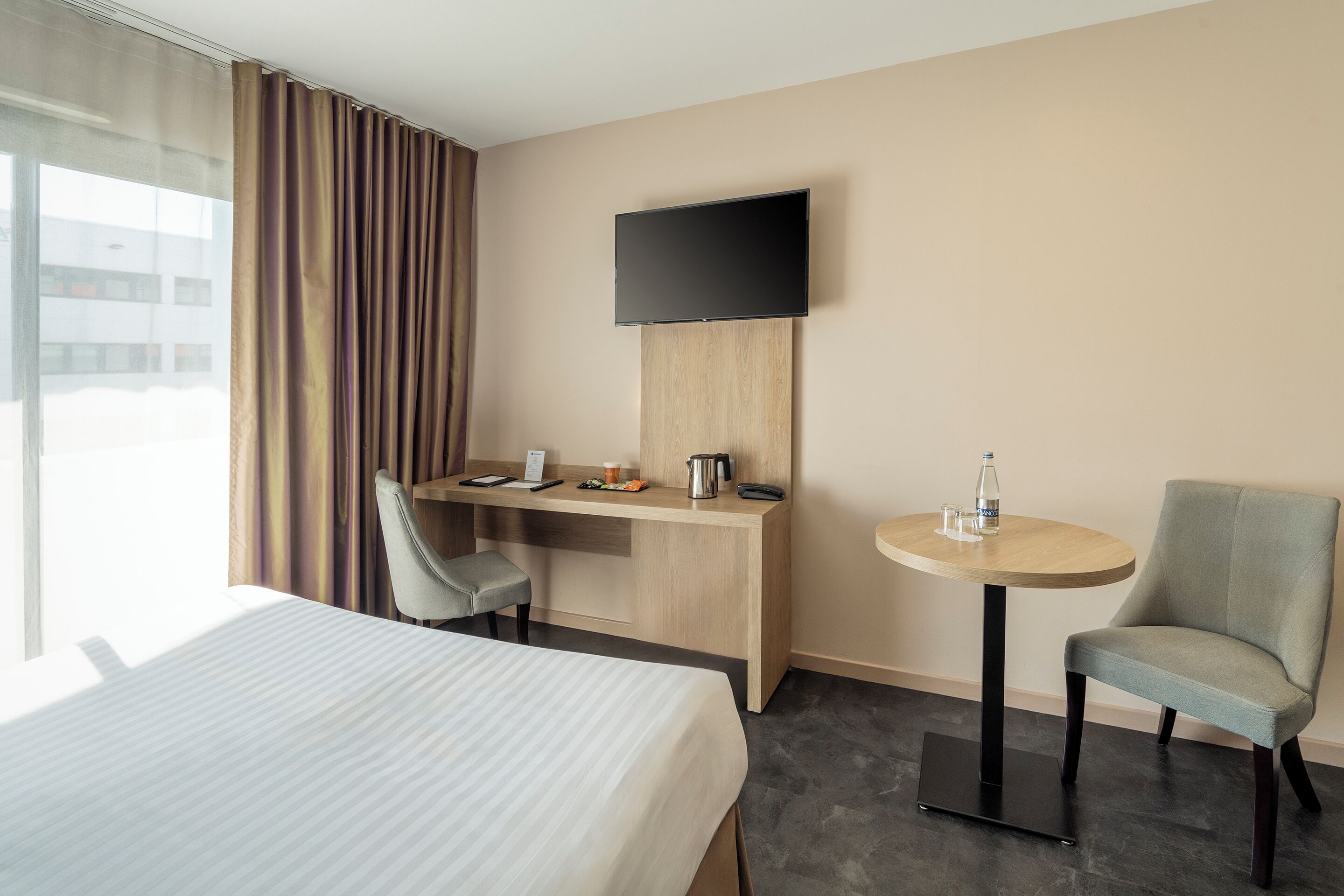 A hotel room with a bed, a desk with a chair, a round table with a chair, a flat-screen TV, and a window with curtains.