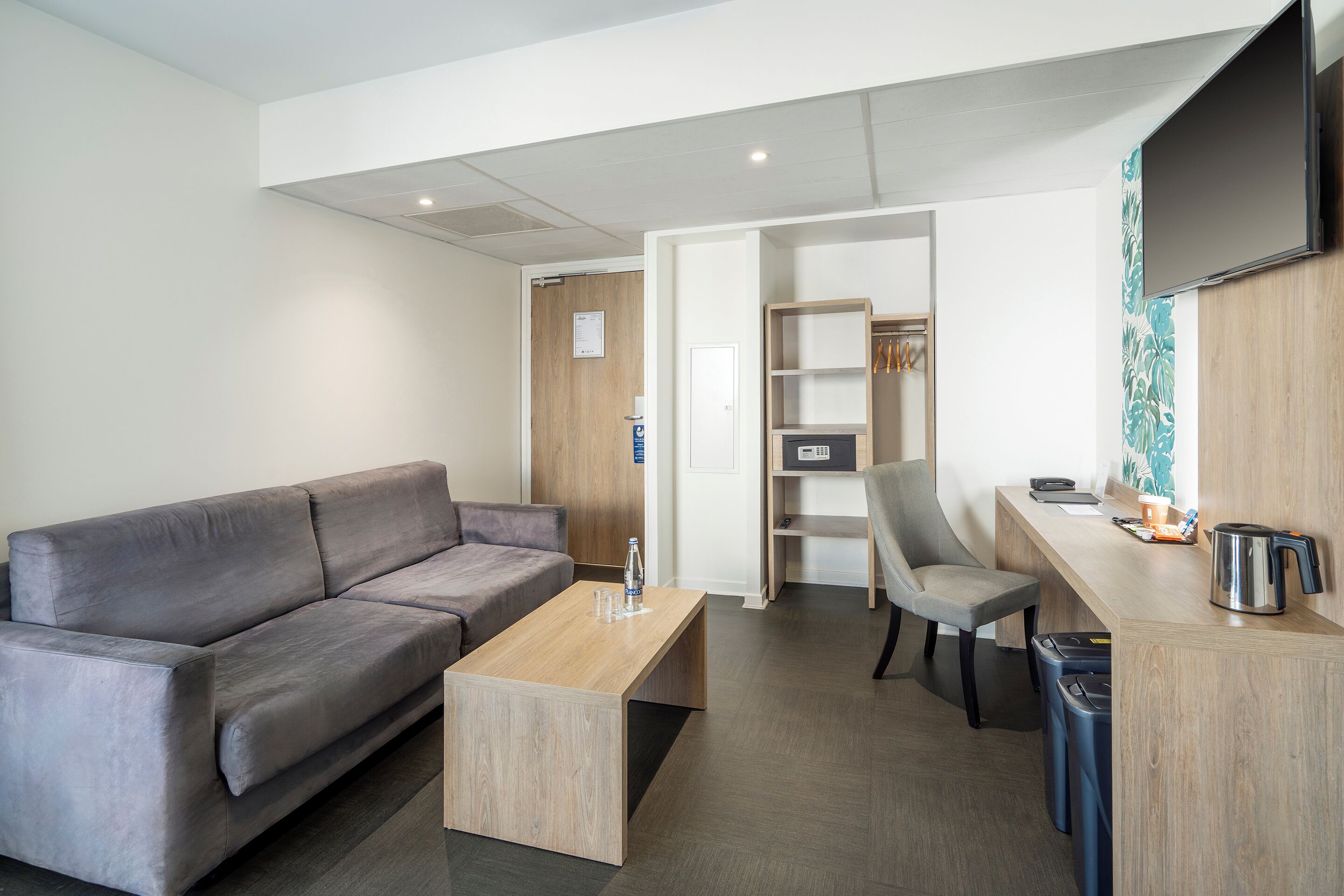 Una sala de estar moderna con un sofá gris, una mesa de centro de madera, un sillón gris, un escritorio pequeño con un hervidor y un televisor montado en la pared.