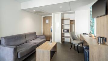 Una sala de estar moderna con un sofá gris, una mesa de centro de madera, un sillón gris, un escritorio pequeño con un hervidor y un televisor montado en la pared.
