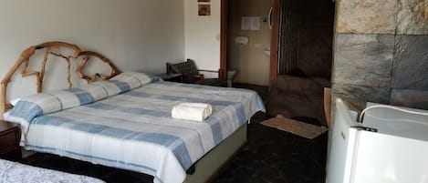Un dormitorio con cama, una silla, una mesita, un refrigerador y una pared de piedra.