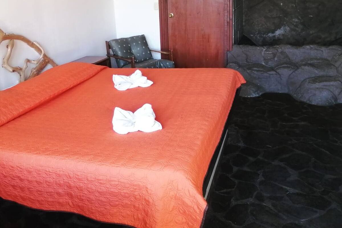 Una cama con una colcha naranja, dos toallas blancas, una silla y una pared de piedra.