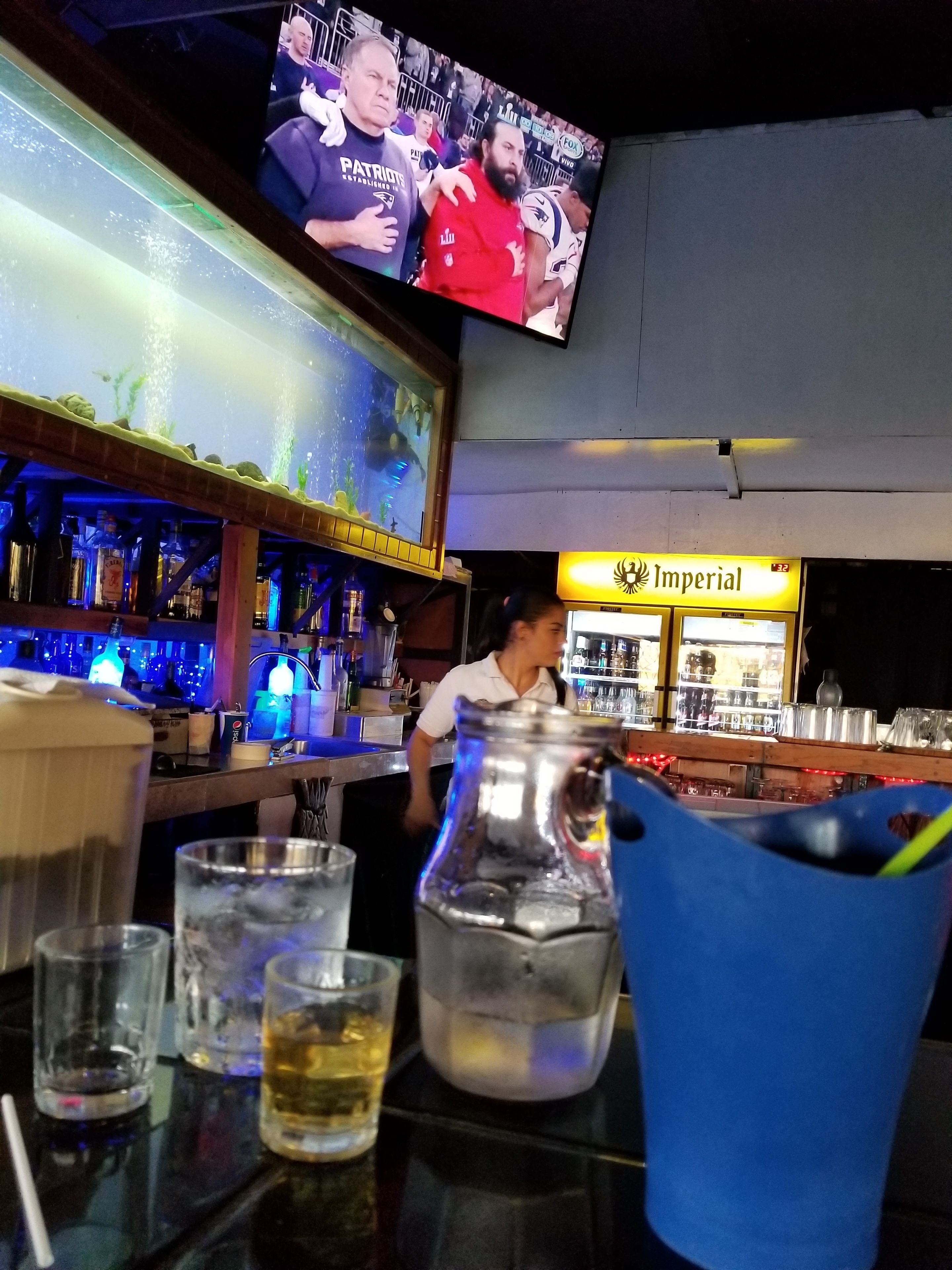 A bar area with a television displaying a sports event, a bartender, and various bar items including glasses and a large pitcher.
