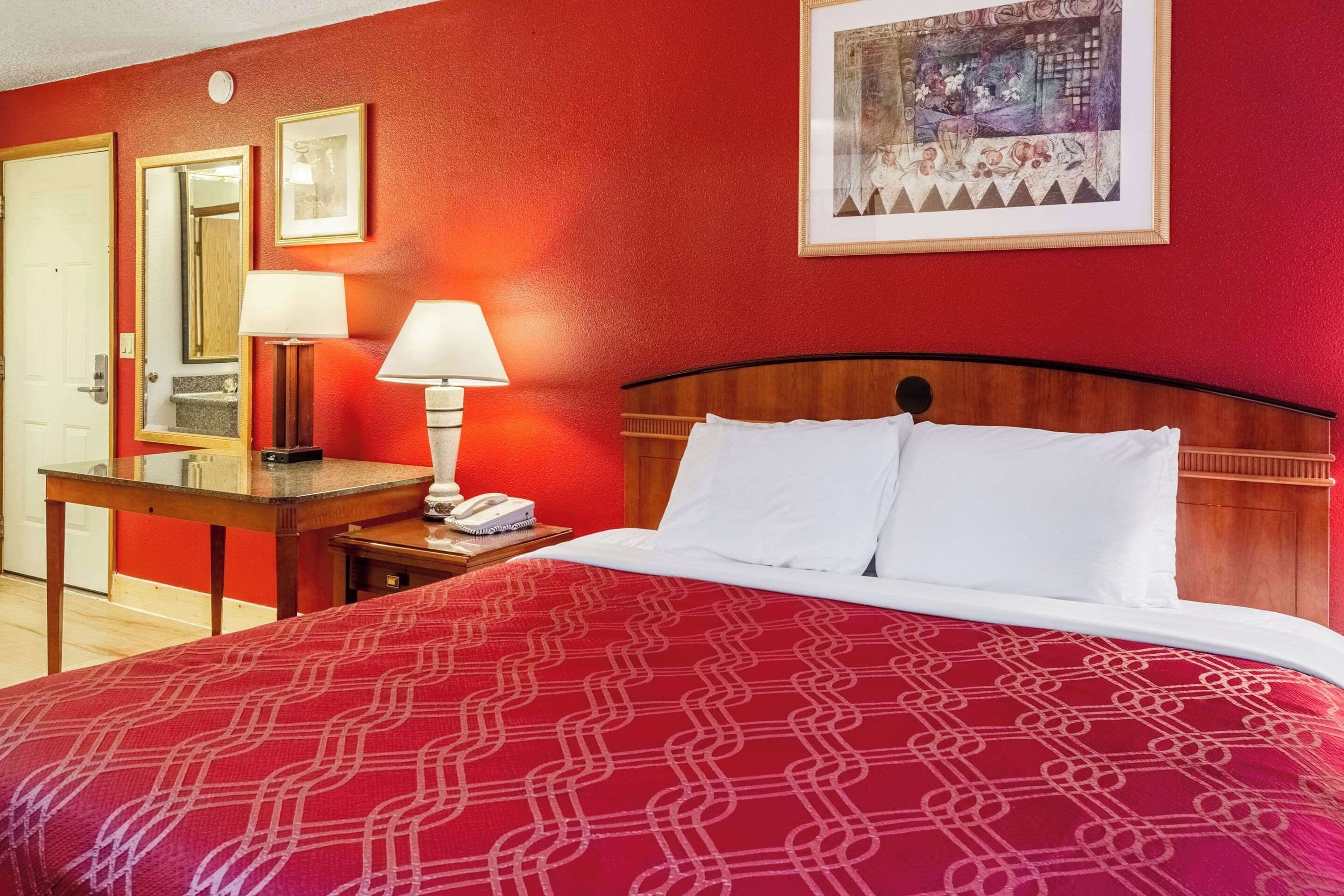 Habitación de hotel con pared roja, estructura de cama de madera, ropa de cama blanca, escritorio con lámpara y teléfono.
