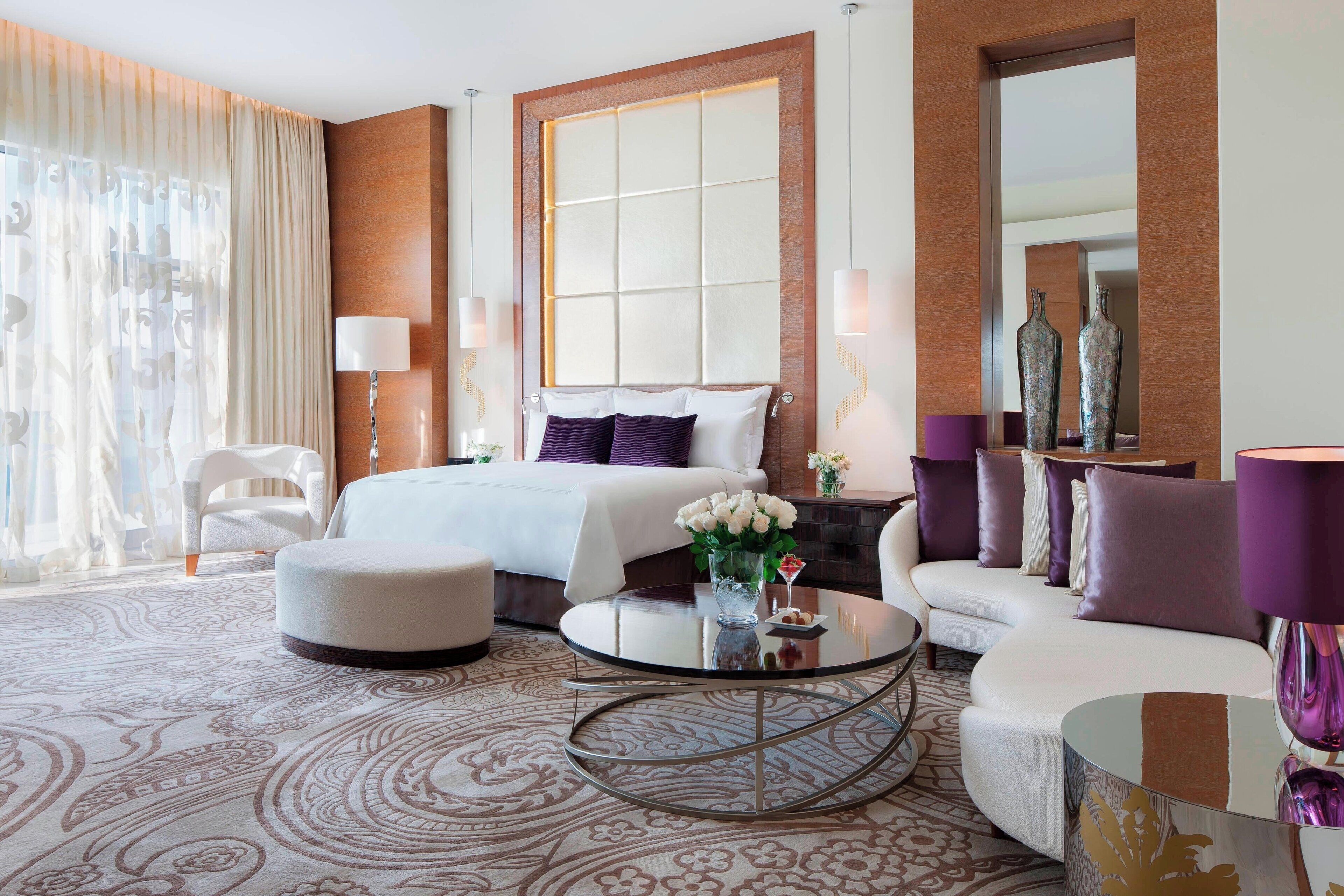 A modern hotel room with a large bed, a sitting area with a sofa and armchairs, a round coffee table, and a large window with curtains.