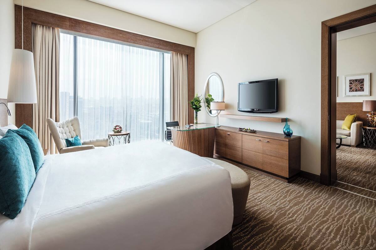 A hotel room with a large bed, a desk with a mirror, a television, and a view of the city through large windows.