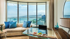 A modern living room with a curved glass coffee table, a beige sofa with blue cushions, and a view of a cityscape through large windows. - JW Marriott Absheron Baku (Baku)
