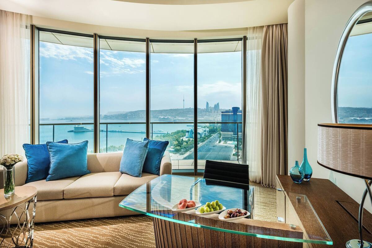 A modern living room with a curved glass coffee table, a beige sofa with blue cushions, and a view of a cityscape through large windows.