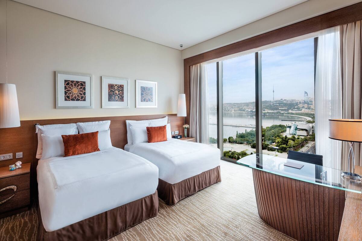 A hotel room with two beds, a glass desk, and a large window offering a city view.