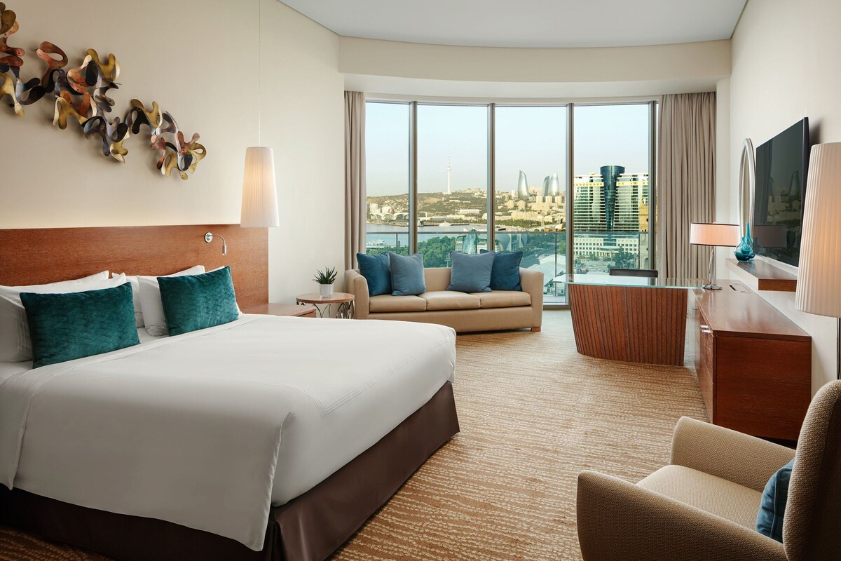 A hotel room with a large bed, a sofa, a desk, and a TV. There is a large window with a view of buildings and a cityscape.