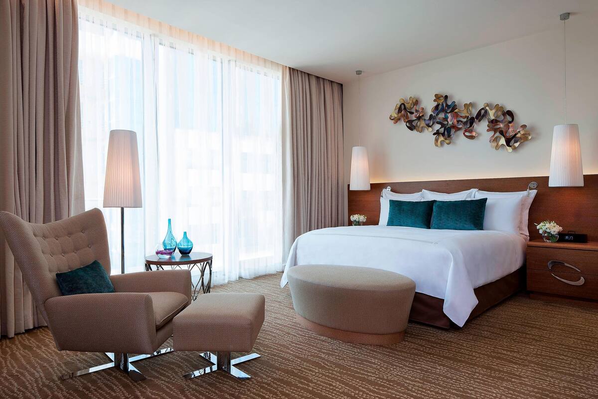 A hotel room with a large bed, a sitting area with a chair and ottoman, a bedside table with a lamp, and a view of the city through large windows.