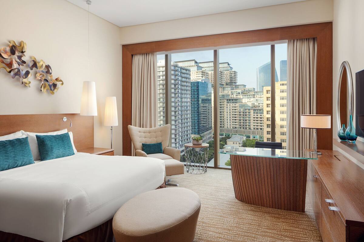 A hotel room with a large bed, a desk, a chair, a view of the cityscape, and a decorative wall piece.