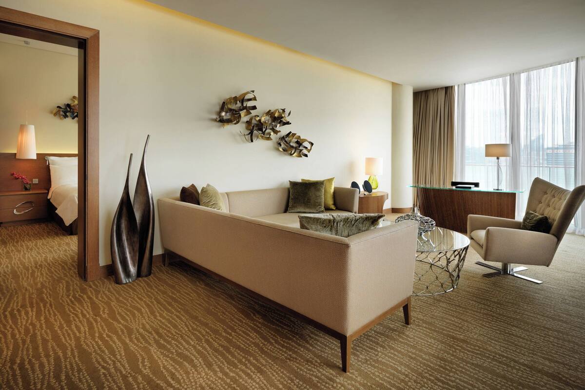 A hotel room with a sofa, armchair, and a glass coffee table. There is a mirror, a lamp, and a decorative wall piece.