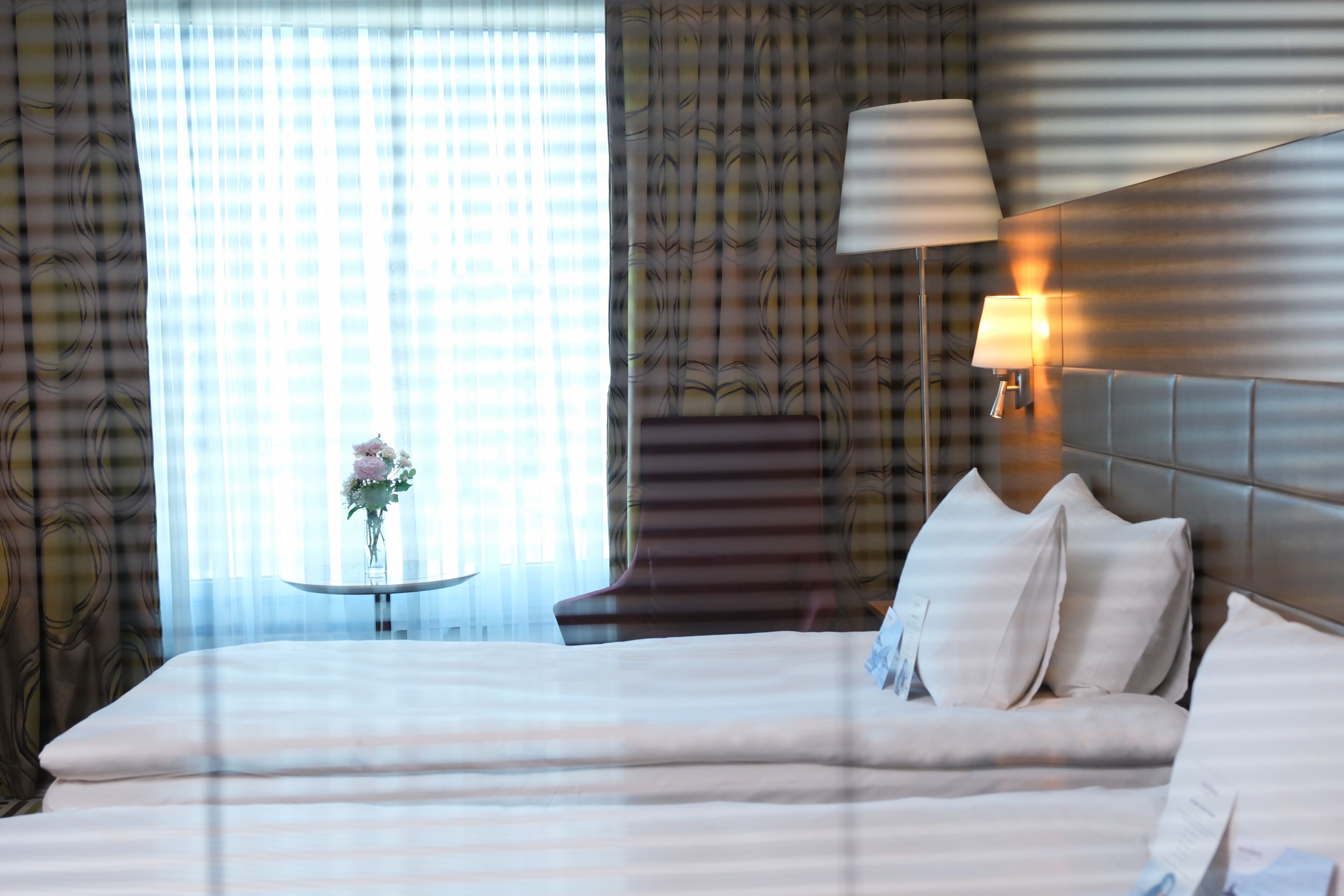 A hotel room with a bed, a bedside lamp, a small table with a vase of flowers, and curtains.