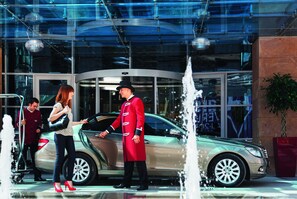 Una entrada de hotel moderna con fachada de vidrio, una fuente y un automóvil estacionado afuera.