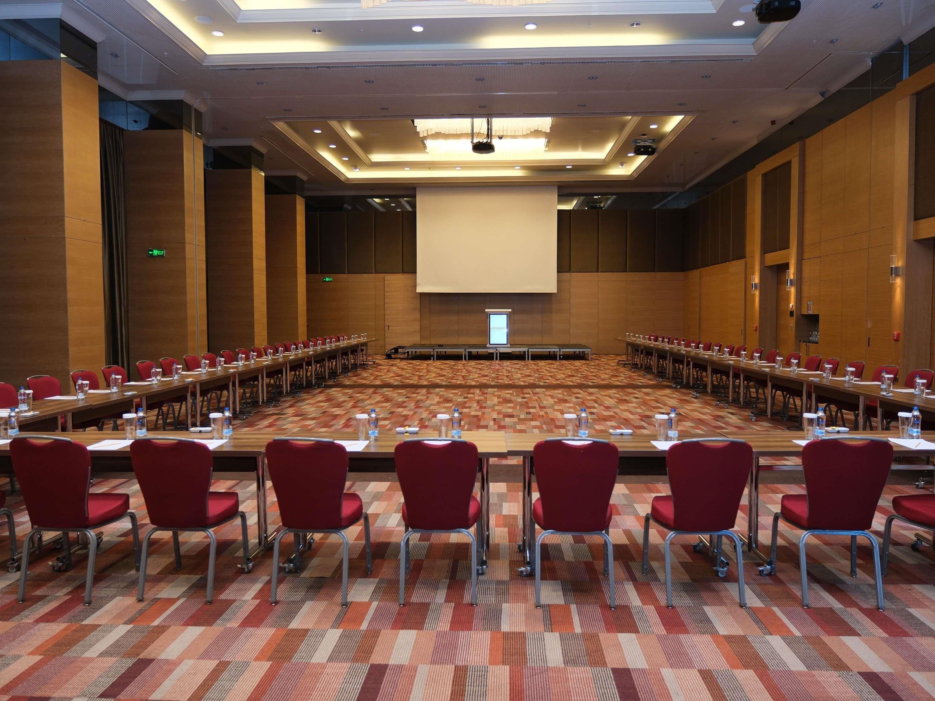 Un salón de conferencias con una larga mesa, sillas rojas y una pantalla grande al frente.