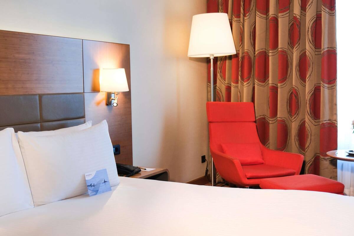 A hotel room with a bed, a red armchair, a nightstand with a lamp, and a patterned curtain.