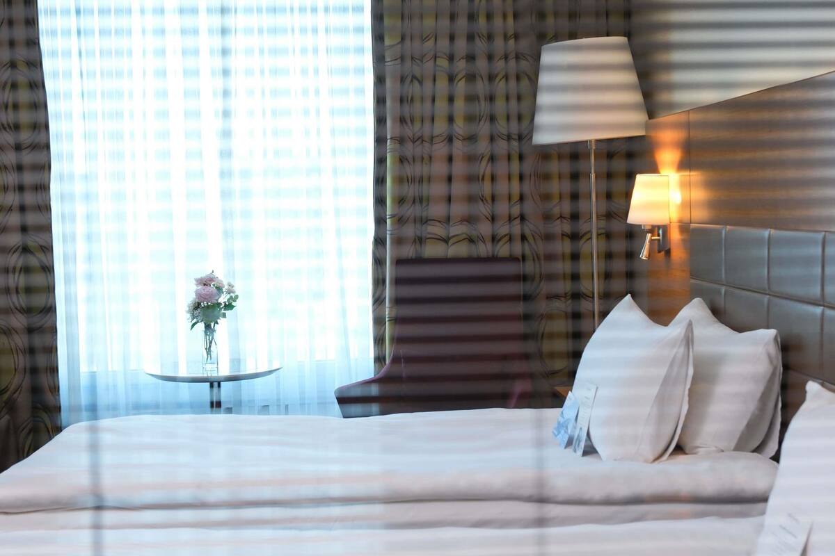 A hotel room with a large bed, a bedside table with a lamp, a window with curtains, and a view of the cityscape.