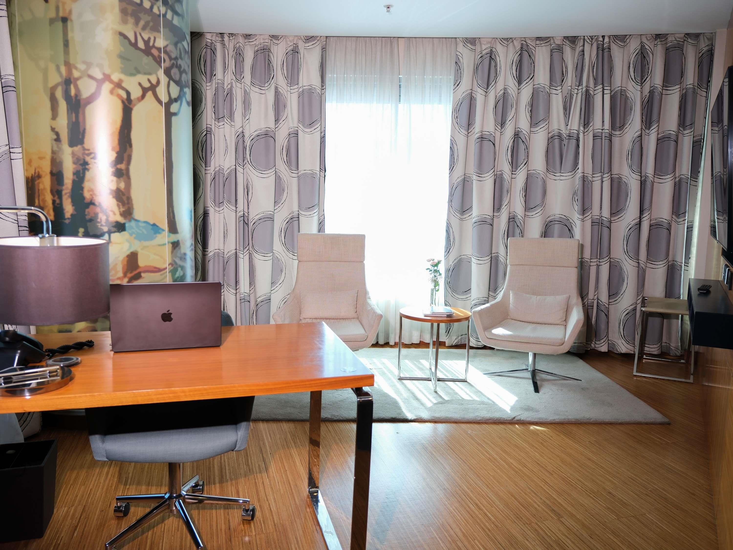 A modern room with a wooden desk, two armchairs, a laptop, a lamp, and a painting on the wall.