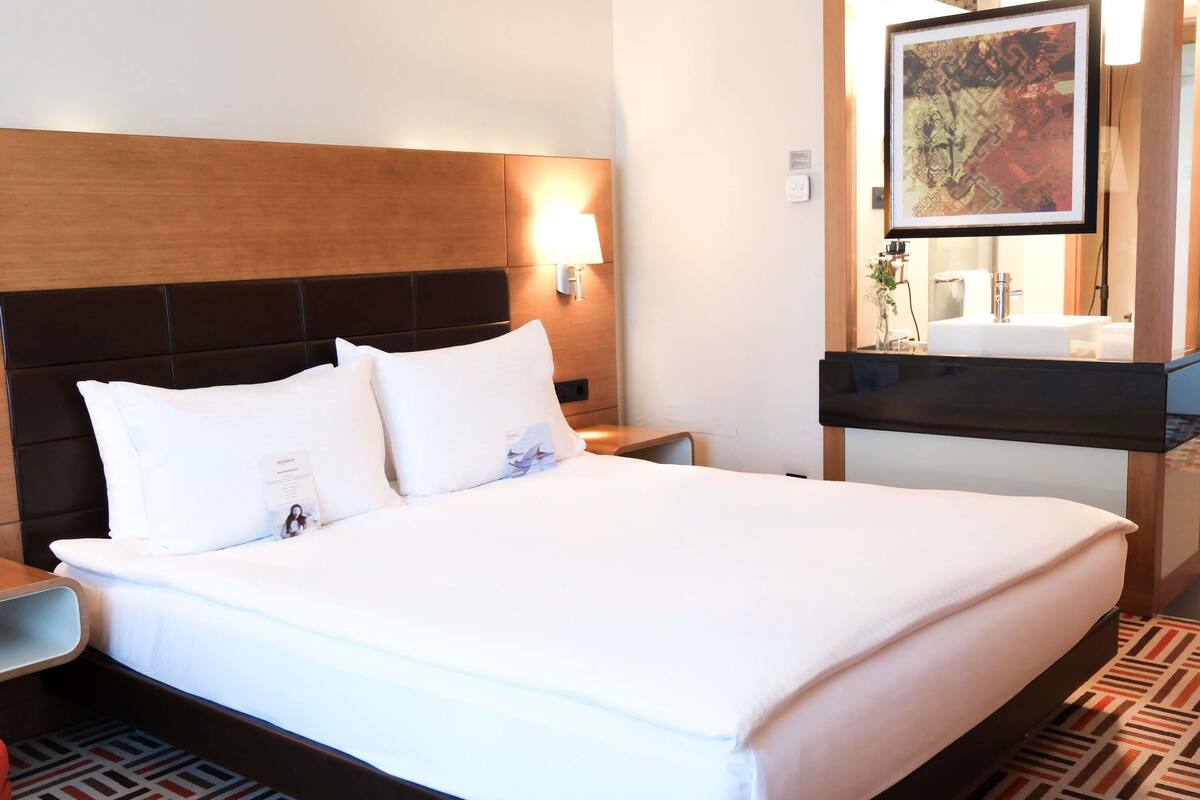 A hotel room with a large bed, a wooden headboard, a bedside table, a nightstand, a wall-mounted lamp, a framed picture, a bathroom with a sink and mirror, and a patterned floor.
